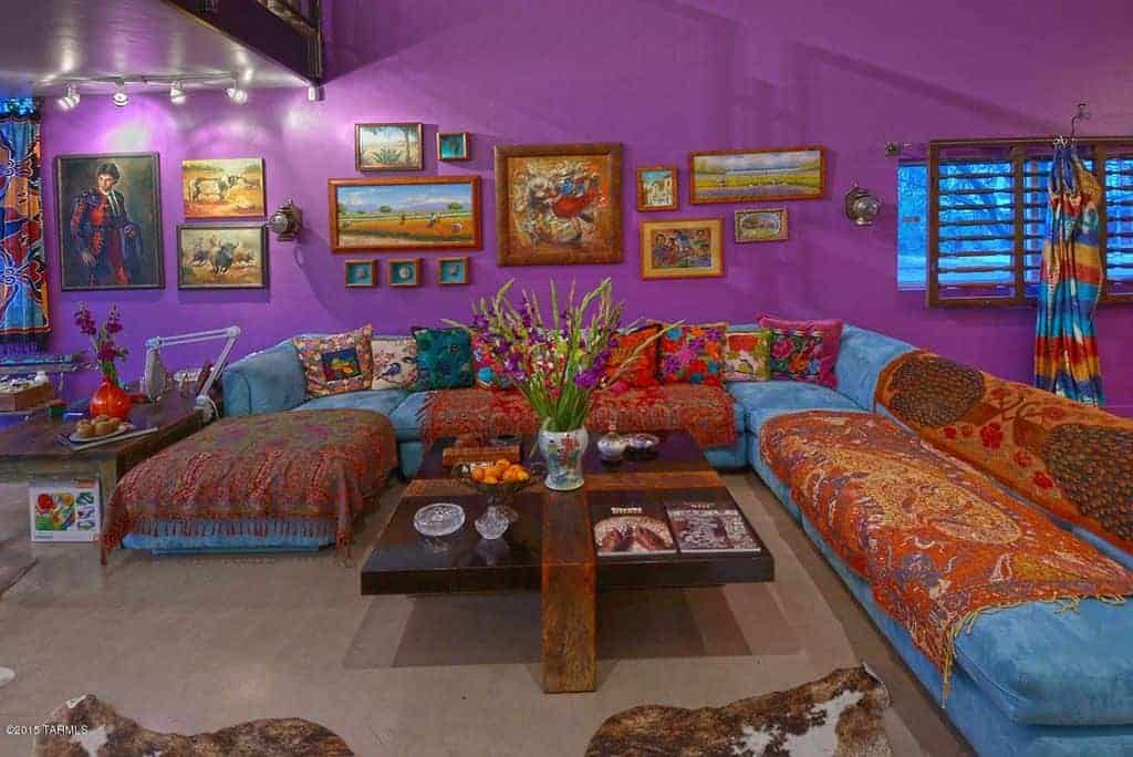 Purple living room designed with gallery frames mounted above the blue velvet sofa. There's a dark wood coffee table in the middle topped with bowls and flower vase.