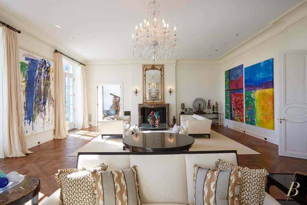 A fancy crystal chandelier illuminates this living room featuring multiple seats and colorful abstract paintings mounted on the white walls. It includes a fireplace and rectangular mirror flanked by candle sconces.