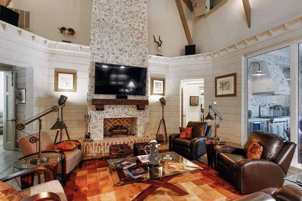 A wall mount TV hangs above the herringbone fireplace fixed on the white textured pillar. This room showcases a glass top coffee table surrounded with cozy seats and round side tables.