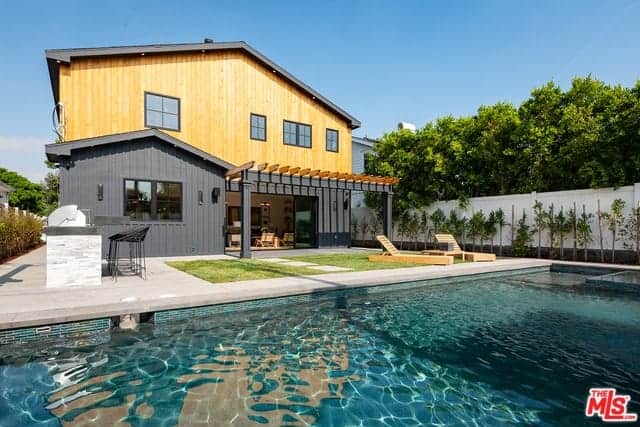 This backyard view showcases the blue pool that mirrors the brilliant blue skies. There are a couple of wooden lawn chairs to enjoy the brilliant view on the small grass lot between the pool and the house that can be accessed through a large entryway under charming trellises.