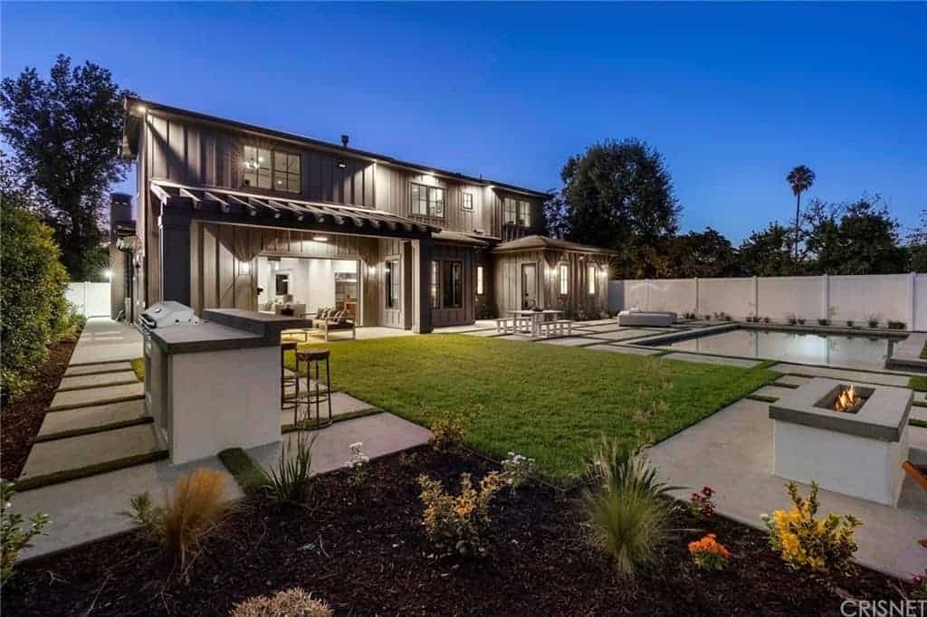 This is a large backyard in a distinct Farmhouse-style landscaping of gray concrete walkways blending with lush green carpets of grass that leads to a pool, an outdoor dining area and the charming firepit that provides warmth for the sitting area.