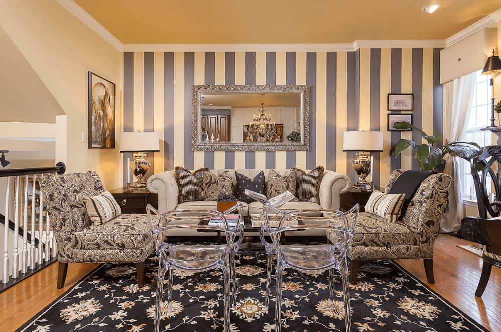 Eclectic living room boasts fabric and glass seats surrounding a black coffee table over a black floral rug. It includes drum table lamps and a gorgeous mirror mounted on the striped accent wall.