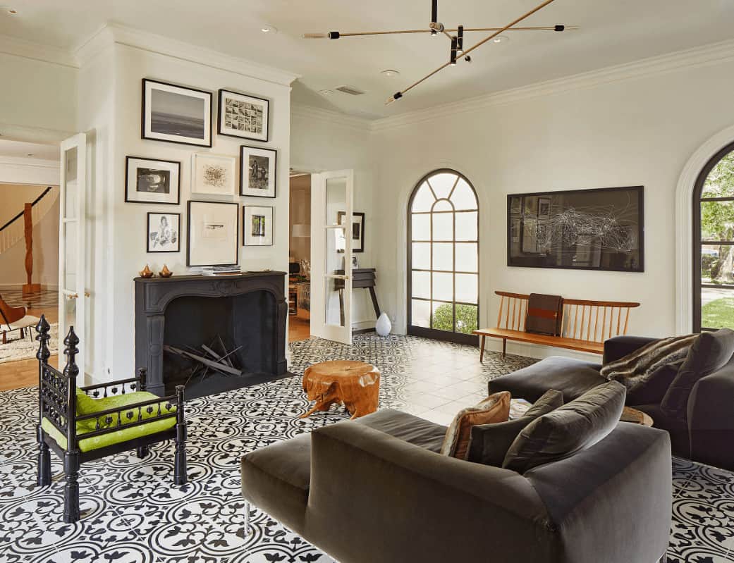 Airy living room with decorative tile flooring and gallery wall art mounted above the black fireplace. It includes velvet chaise lounges and a green cushioned chair paired with a stump coffee table.
