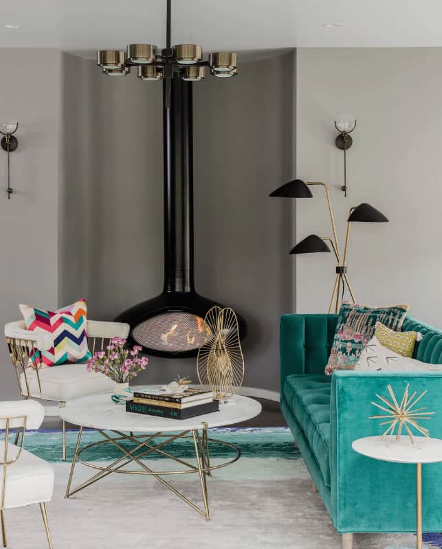 This living room boasts a green velvet sofa and white round back chairs surrounding a modern coffee table. It includes a stylish chandelier and black hanging fireplace that complements the three head floor lamp.