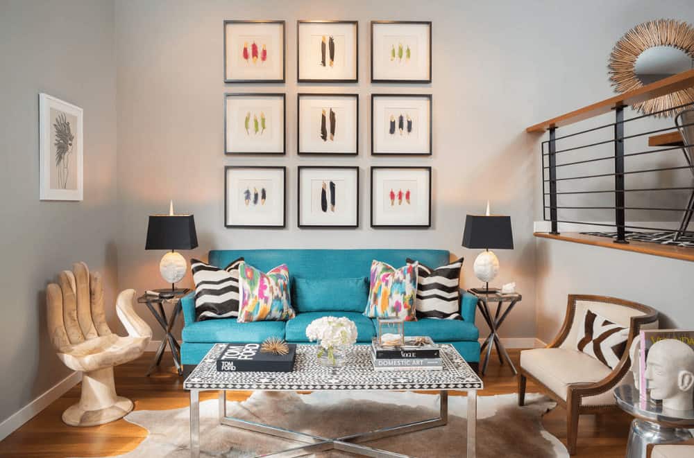 A feather art gallery sets a gorgeous backdrop to the blue sectional sofa accented with multi-colored and black and white pillows. It is accompanied by hand and beige chairs along with a metal coffee table on a cowhide rug.