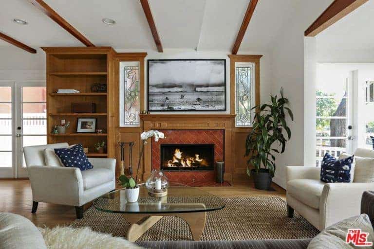 Cottage style living room boasts a fireplace with black and white artwork on top along with a glass top coffee table accompanied by white cushioned chairs accented with deep blue pillows.