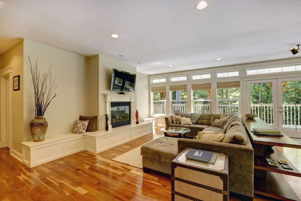 A flat-screen TV hangs above the fireplace facing the velvet tufted sofa and round glass top coffee table over a beige area rug. This room has rich hardwood flooring along with glass paneled windows and French door leading out to the balcony.