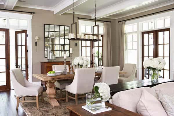 The white coffered ceiling of this dining area beside the living room supports a decorative lighting over the wooden dining table. This looks like a tray filled with candles that matches the candelabras on the dining table that is surrounded with white cushioned chairs.