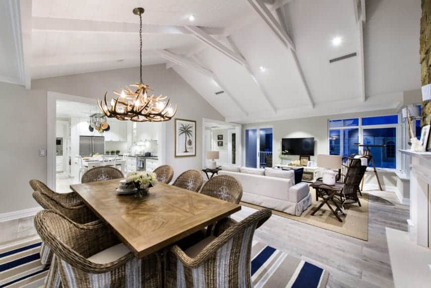 This great room that houses both the dining area and the living room area has a high white cathedral ceiling with exposed beams. It hangs a majestic antler chandelier over the wooden dining table that is partnered with woven wicker chairs.