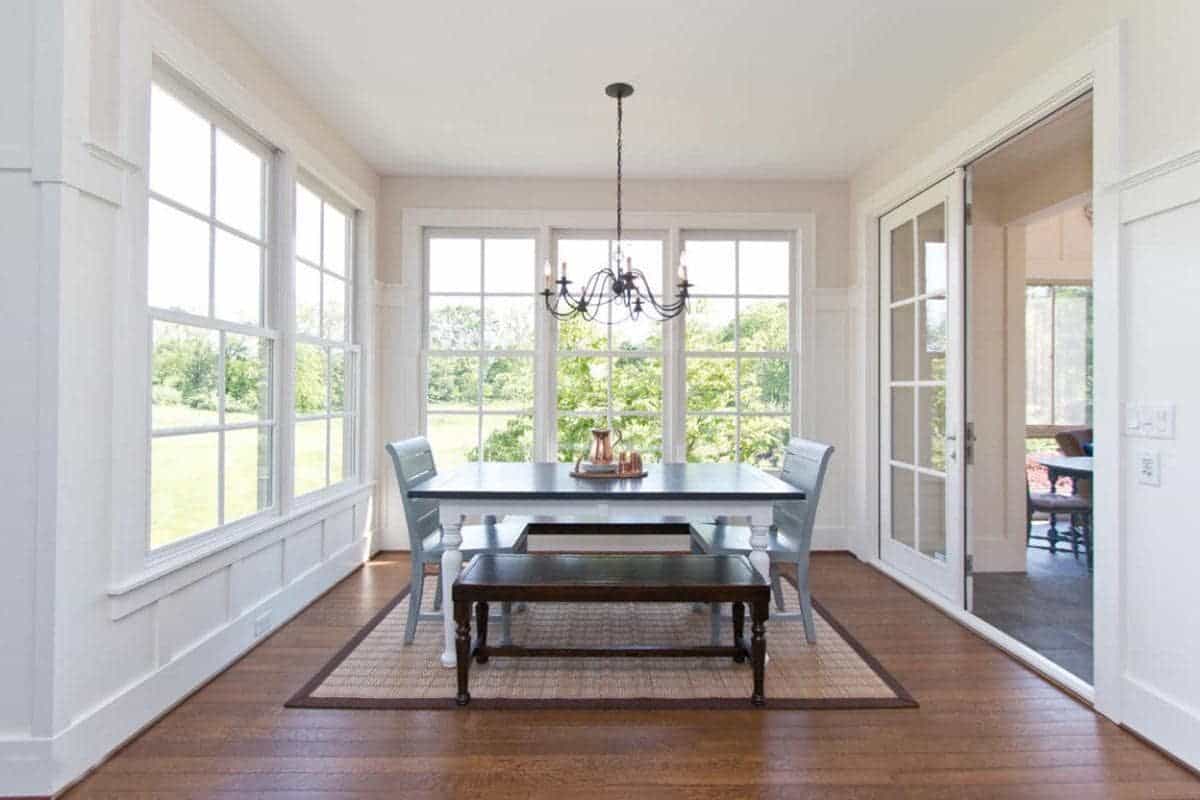 The highlight of this Farmhouse-style dining room is the lush green scenery outside the tall French windows that dominate the white walls contrasted by the dark hardwood flooring that pairs well with the wooden dining table and its wooden benches.