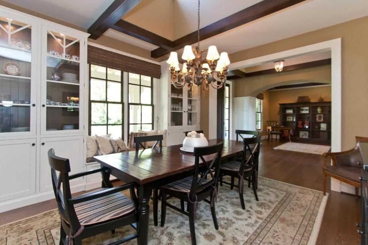 The beige ceiling has wooden exposed beams that have a dark brown hue matching with the hues of the Farmhouse-style wooden dining set over the beige patterned area rug of the dark hardwood flooring. These are all augmented by the yellow lights of the chandelier.