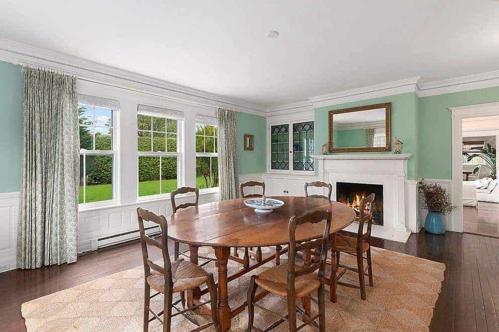 This Farmhouse-style dining room has a simple wooden dining set with wooden chairs that have woven wicker seats that match the area rug over the dark hardwood flooring. This setup is warmed by the fireplace by the head of the table. This has a white mantle that stands out against the green walls.