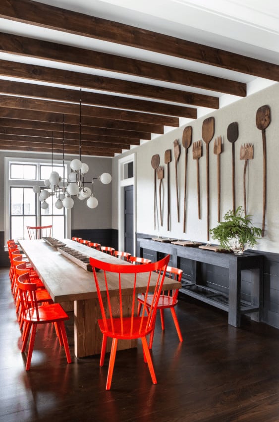 This dining room has a white ceiling dominated by the exposed wooden beams that match the hardwood flooring and the large rectangular wooden dining table surrounded by brilliant red orange wooden chairs and topped with a peculiar modern chandelier.