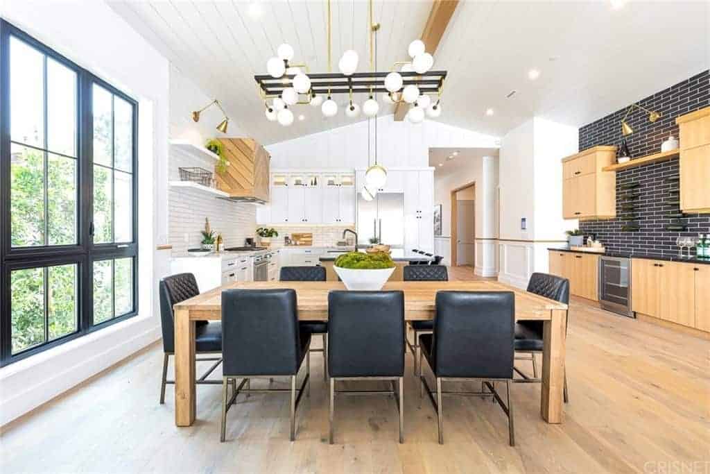 The black leather cushions of the modern dining chairs matches with the black frames of the large window, the black brick backsplash of the kitchen and the black metal frames of the modern lighting above the simple wooden dining table.