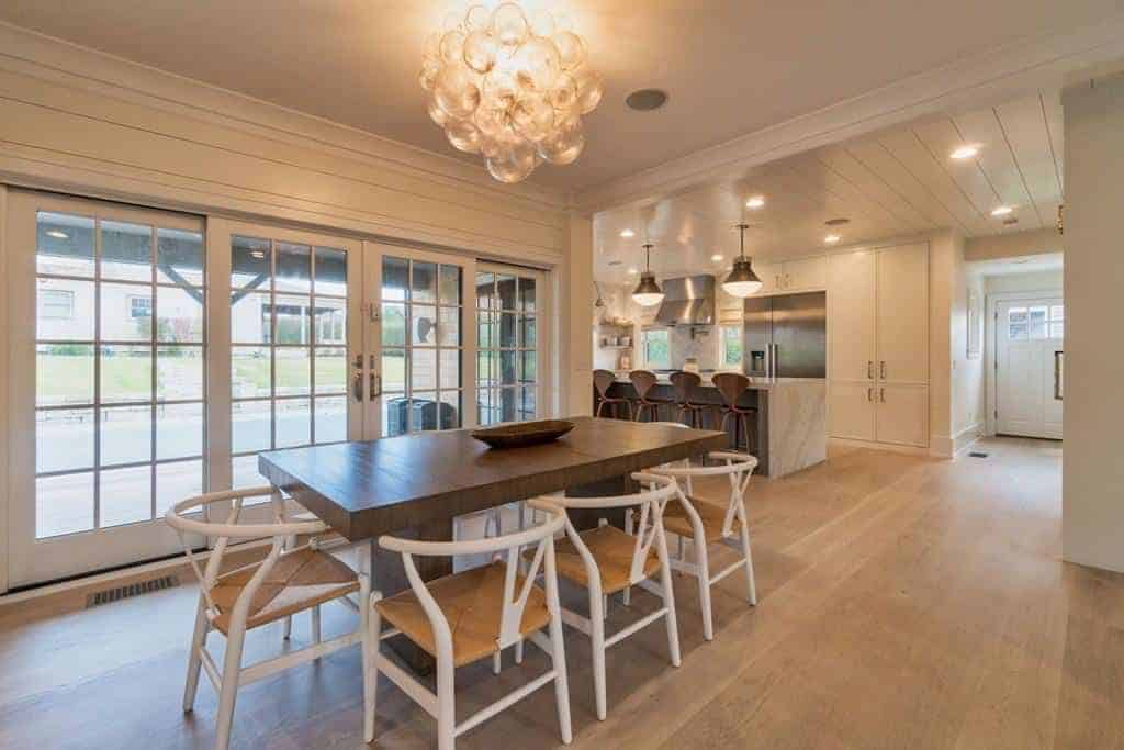 This Farmhouse-style dining room has white wooden wishbone chairs with rustic woven wicker seats that complement the hardwood flooring and the rectangular wooden dining table that has a slightly darker brown hue warmed by the yellow lights of the decorative lighting above.