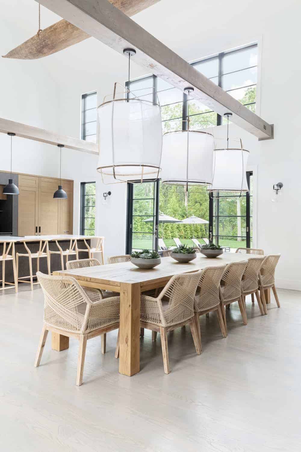 The high ceiling is paired with tall white walls with tall windows that bring in an abundance of natural lighting that complements the wooden dining table paired with rustic woven wicker armchairs. This is topped with a large floating beam supporting hooded pendant lights.