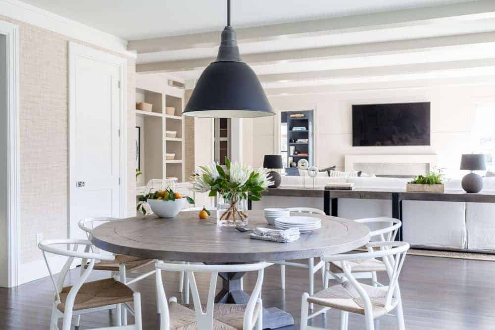 The round wooden dining table blends with the dark hardwood flooring that contrasts the white wooden wishbone chairs with woven wicker seats. This charming aesthetic is capped off with a farmhouse-style dome pendant light hanging from the white ceiling.
