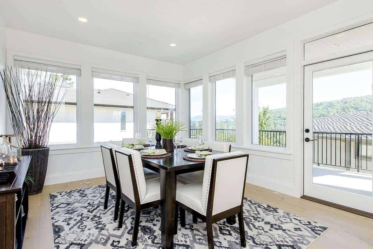 The white brightness of this Farmhouse-style dining room is due to the surrounding glass windows and glass door that bring in natural lighting to the white ceiling with recessed lights and the white walls that match the white leather upholstery of the chairs.