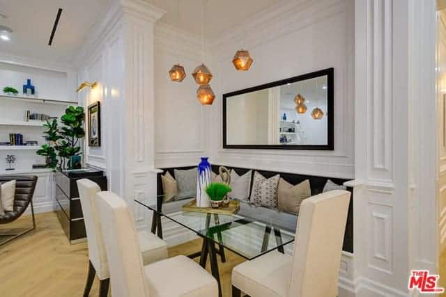This is an informal dining area by the living room area. This is embedded into a tiny alcove of the white wall that has a built-in bench with black cushions paired with a rectangular glass-top dining table and beige cushioned chairs with black wooden legs.