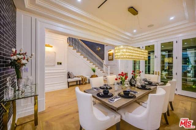 This elegant Farmhouse-style dining room has a white tray ceiling that supports a brilliant golden decorative lighting over the wooden dining table that is paired with white cushioned chairs that contrast the black wall with brick wall patterns.