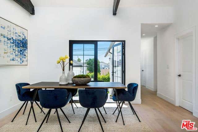 The blue velvet cushioned chairs are the highlights of this simple Farmhouse-style dining room. It matches with the colorful abstract painting mounted on the white wall by the head of the wooden dining table that complements the hardwood flooring.