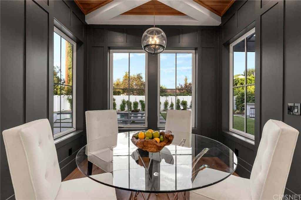 This informal dining area is inside a small nook alcove of dark gray walls with windows that bring in natural lighting for the round glass-top table and its white cushioned chairs that pairs well with the white exposed beams of the wooden ceiling.