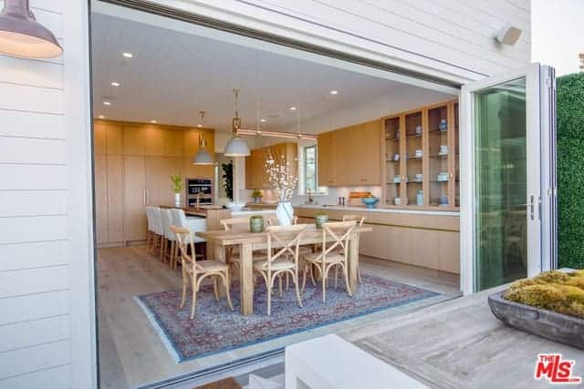 This is an informal Farmhouse-style dining area by the kitchen. The rustic wooden dining set is bathed in natural lights coming from the entire wall opening up courtesy of the folding glass doors with green tint and white frames that match the white ceiling.