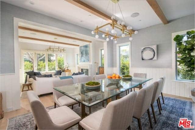 The glass-top dining table is paired with light gray cushioned dining chairs that stand out against the blue patterned area rug underneath. This dining room has a white ceiling with exposed wooden beams and a golden modern chandelier.