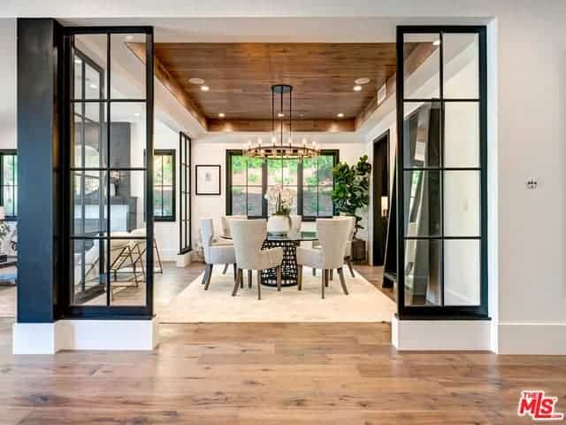 This is an elegant Farmhouse-style dining room with a charming wooden tray ceiling that supports a round Farmhouse-style chandelier complementing the round glass-top dining table surrounded by cushioned wing-back chairs.