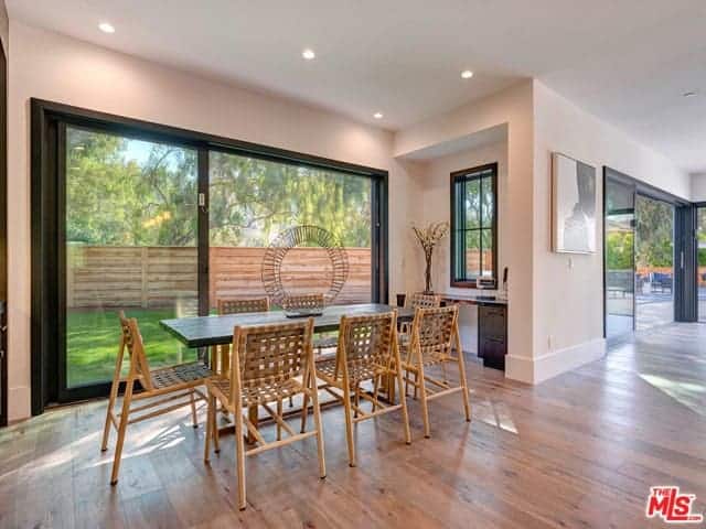 This simple dining room has a beautiful lush green background of the landscaping outside with a brown wooden fence. This scenery complements the rustic wooden dining chairs that have woven backs and seats matching with the hardwood flooring.