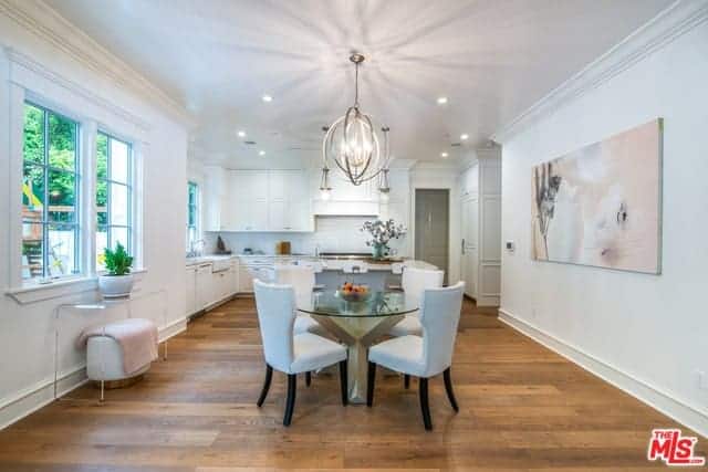 This small dining area has a stainless steel spherical decorative lighting hanging over the round glass-top dining table and its white cushioned wing-back chairs that stand out against the hardwood flooring but blends with the white walls.