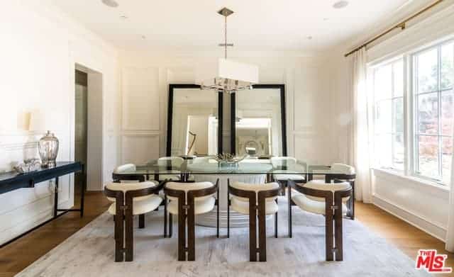 The white walls and ceiling of this Farmhouse-style dining room are further brightened by the ample natural lighting coming from the large curtained window by the head of the rectangular glass-top dining table paired with modern chairs.