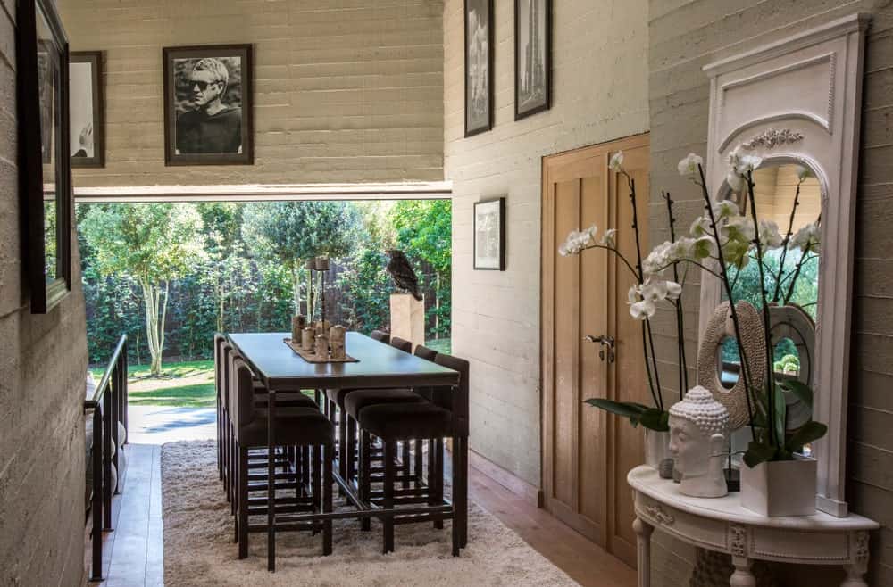 This simple dining area has a background of lush green landscapes outside of the open wall by the head of the rectangular dark wooden dining table that is paired with wooden dining stools with dark cushions contrasting the white furry area rug.