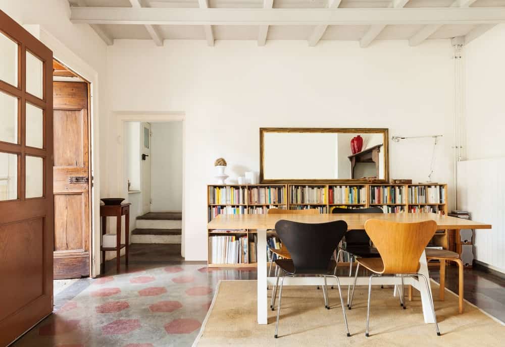 The white wooden ceiling of this Farmhouse-style dining room has white wooden exposed beams that blend with the white walls that are adorned with a large mirror on a row of low bookshelves beside the wooden dining table and its modern chairs.