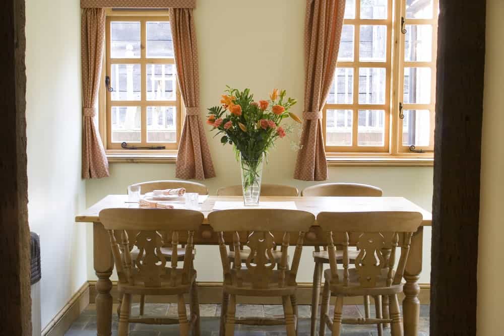 The charming wooden dining table has a simple design to its wooden legs that match with the designs and finish of the wooden dining chairs. This setup is given a nice background of brilliant windows with pink patterned curtains.