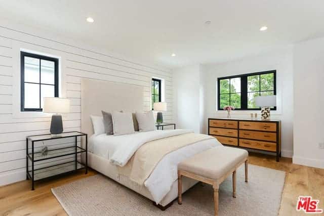 This is a simple and bright Farmhouse-style bedroom with white walls and white ceiling with recessed lights. These are further lightened by the natural lights coming in from the windows flanking the beige cushioned bed with white sheets.