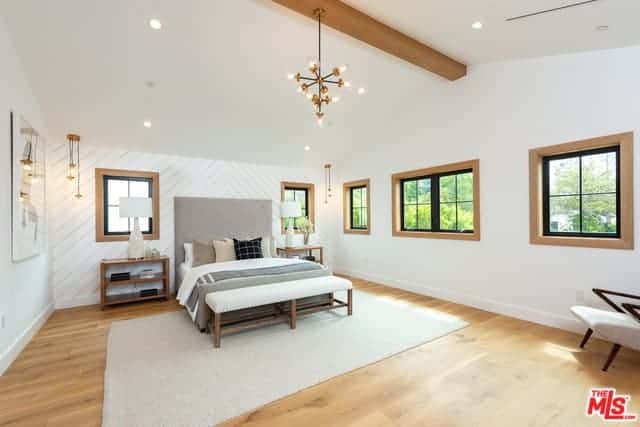 This is a bright and spacious Farmhouse-style bedroom that has a cathedral with a single wooden exposed beam that matches with the hardwood flooring and the frames of the surrounding windows that brighten up the white walls and gray traditional bed.