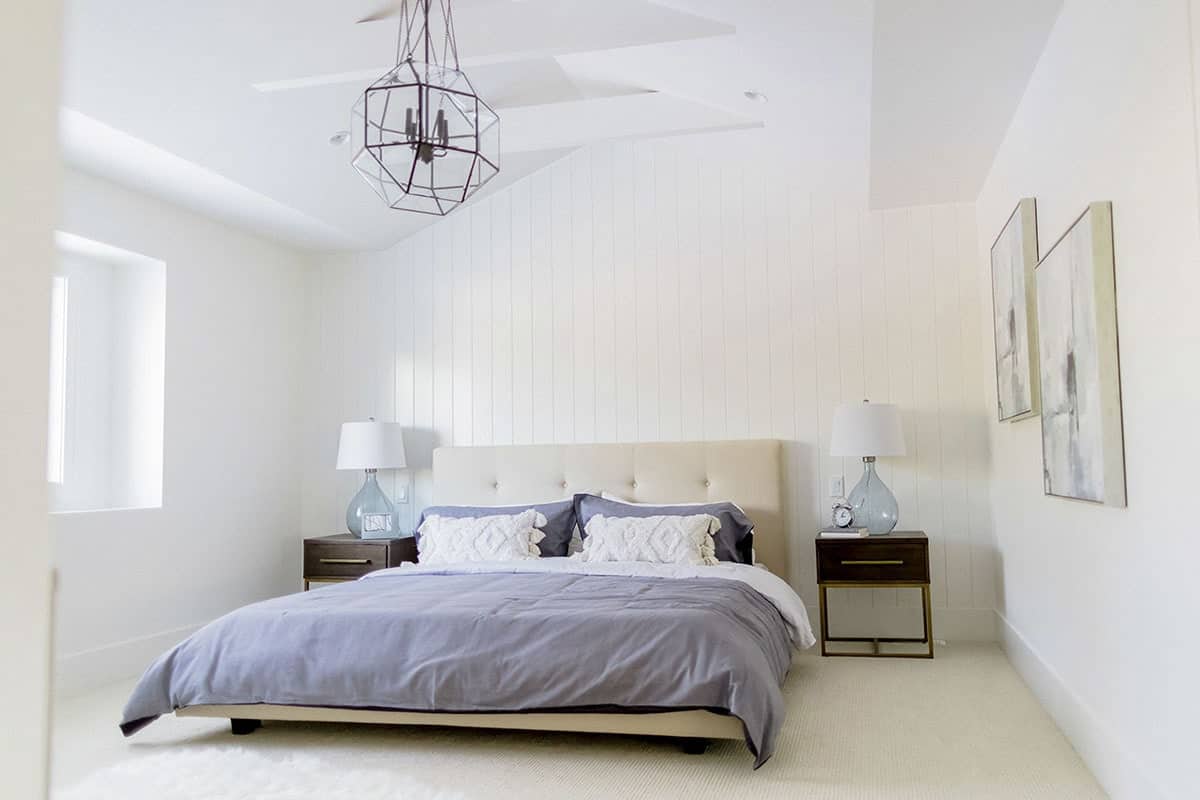 This low platform bed has a beige tufted cushioned headboard that complements the white wall behind it that has a shiplap plank finish. This bed is flanked by dark wooden bedside drawers that contrast the beige carpeted flooring.