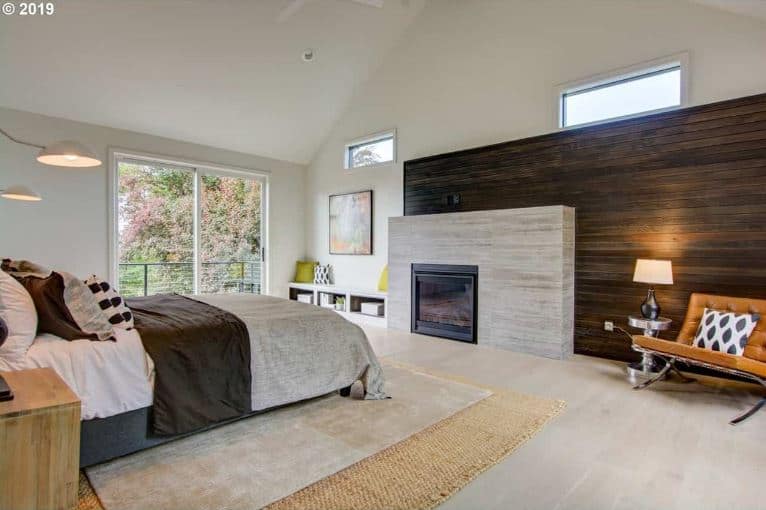 The light hardwood flooring of this elegant Farmhouse-style bedroom is topped with a rustic woven area rug paired with a gray area rug on top of it. This is a perfect match for the concrete slab that houses the modern fireplace by the foot of the bed. This concrete slab is adorned with a large wooden wall with a plank finish.