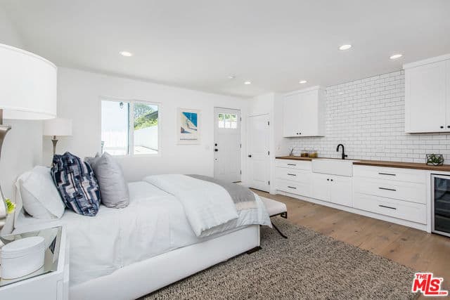 This bedroom has a peculiar design to its setup. The white bed with white sheets blends with the white walls and flanked by table lamps. Across from it is a kitchen peninsula with a faucet and sink with a backsplash of white tiles arranged in a brick wall pattern.