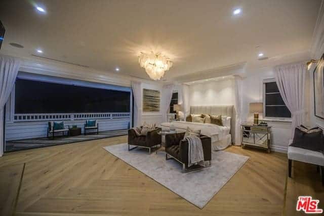 This is a large primary bedroom that has a Farmhouse-style wide hardwood flooring and a large wall that opens up to a balcony. The wide beige ceiling has recessed lights on the sides and the a large decorative lighting in the middle over the sitting area by the foot of the bed.