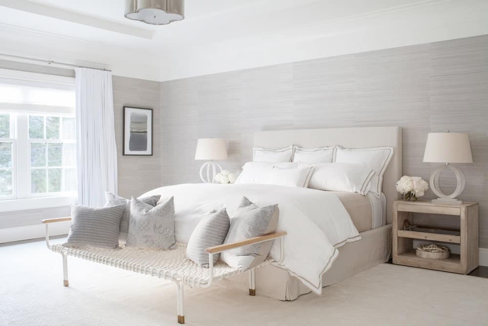 This chic and bright Farmhouse-style bedroom has a beige cushioned bed flanked with wooden bedside tables bearing table lamps with matching beige hoods. This is complemented by the light gray walls and white tray ceiling brightened by the window.