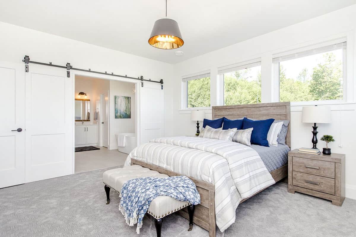 The light gray carpeted flooring of this Farmhouse-style bedroom is a perfect complement to the wooden sleigh bed with a wooden headboard and its matching bedside drawers with the same wooden hue that complements the white walls.