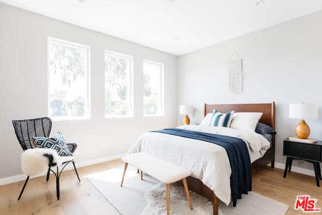 This bright bedroom has white walls and a white ceiling that are brightened even further by the natural lights coming in from the row of windows on the side of the dark wooden bed. This is counterbalanced by the hardwood flooring and the black wooden bedside drawers of the dark wooden bed.