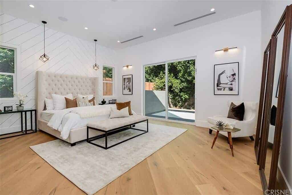 The white wall behind the beige tufted headboard has a wooden plank finish that goes well with the hardwood flooring of this Farmhouse-style bedroom adorned with wall-mounted paintings and large wood-framed mirrors leaning on the wall.