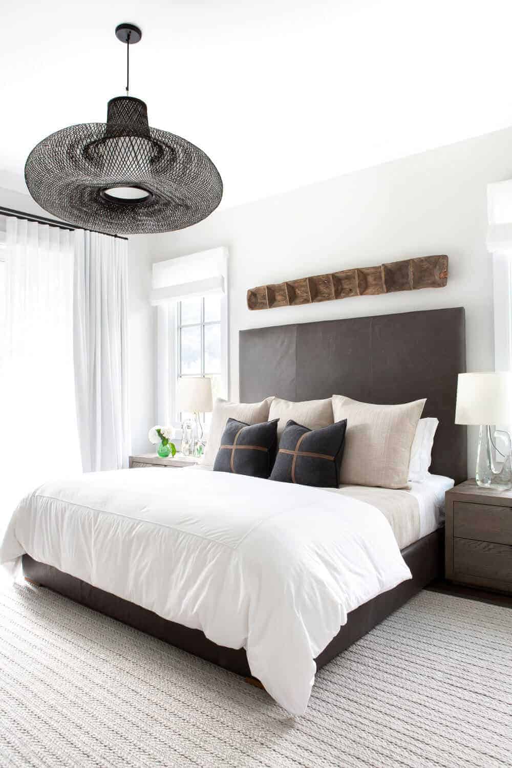 The light gray carpeted flooring complements the dark gray cushioned bed with a large cushioned headboard of the same hue. This is adorned with a wooden artwork mounted on the white wall above the headboard that matches with the decorative hood of the pendant light.