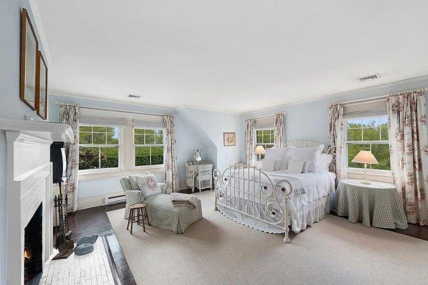 This beautiful Farmhouse-style bedroom has bright blue walls that are complemented by the white ceiling and the natural lights coming in from the windows. This is mirrored by the white wrought iron bed across from the white mantle of the fireplace.