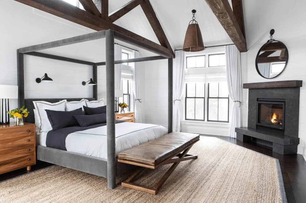 This Farmhouse-style bedroom has a gray four-poster bed that matches the fireplace in the corner with a dark gray mantle that stands out against the white walls. This is also contrasted by the large wooden beams of the white cathedral ceiling.