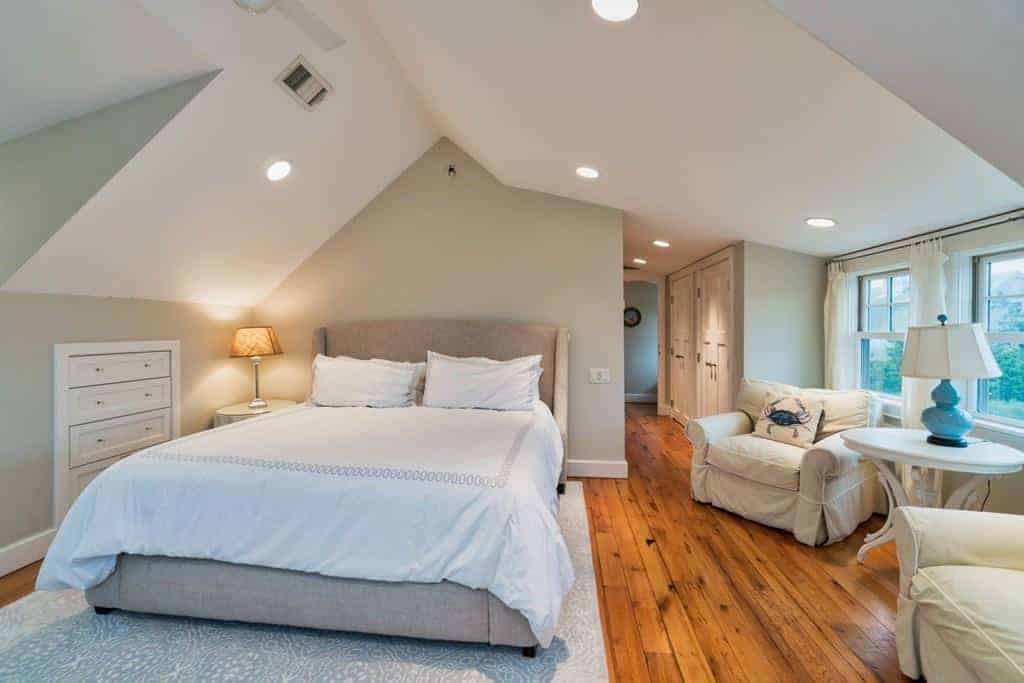 This comfortable bedroom has an irregular arch to its white ceiling that has multiple recessed lights. This is complemented by the light gray walls and contrasted by the hardwood flooring. There is a comfortable sitting area beside the bed that has a couple of cushioned beige armchairs.