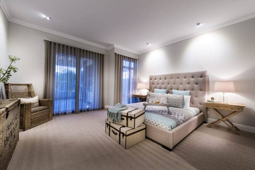 This simple and comfortable bedroom that has a beige carpeted flooring is adorned with various rustic elements like the woven wicker armchair at the corner, the wicker basket that serves as a console table and the pair of wooden bedside tables flanking the beige headboard.